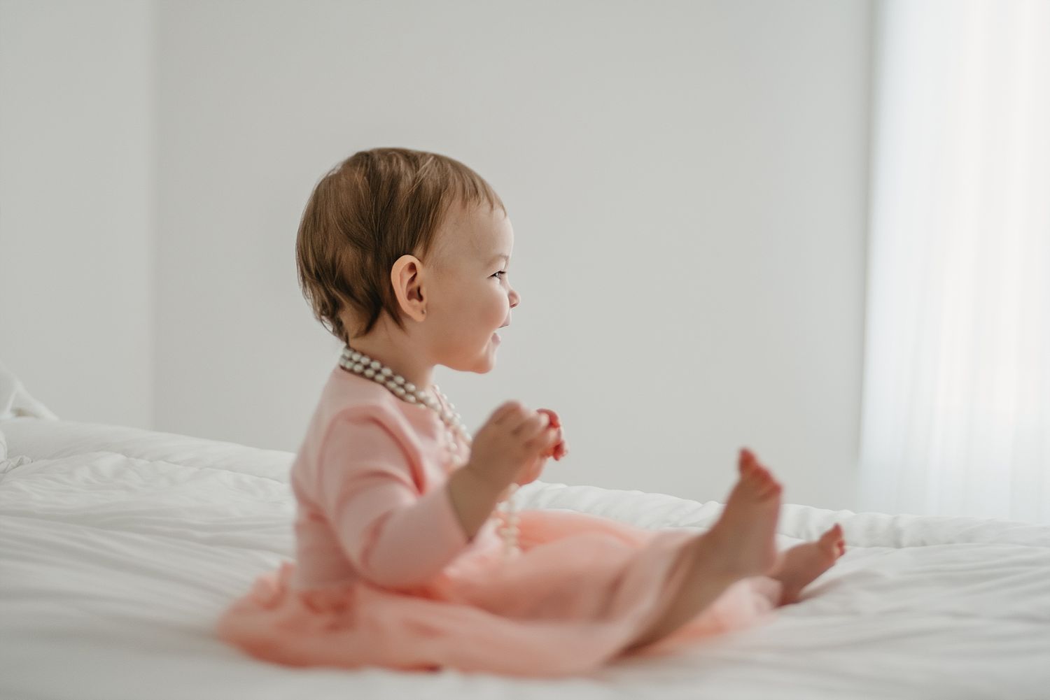 A toddler in pink sits on white bedding in a bright room.