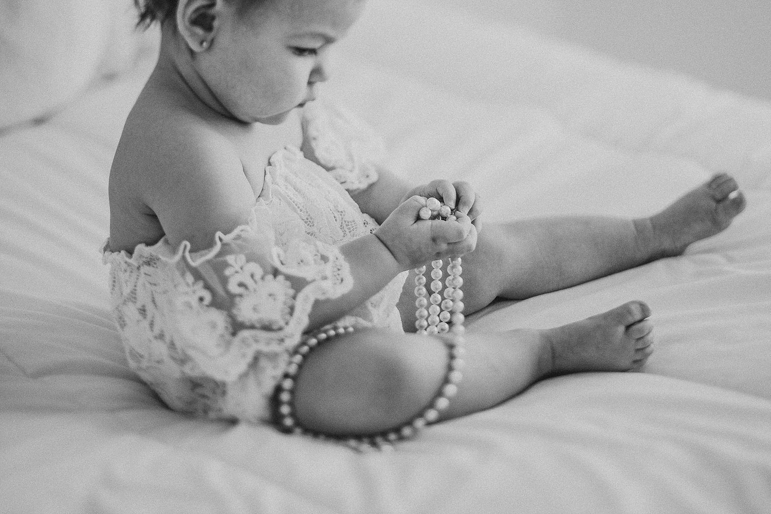 Baby wearing lace outfit plays with pearl necklace while sitting on white bedding.