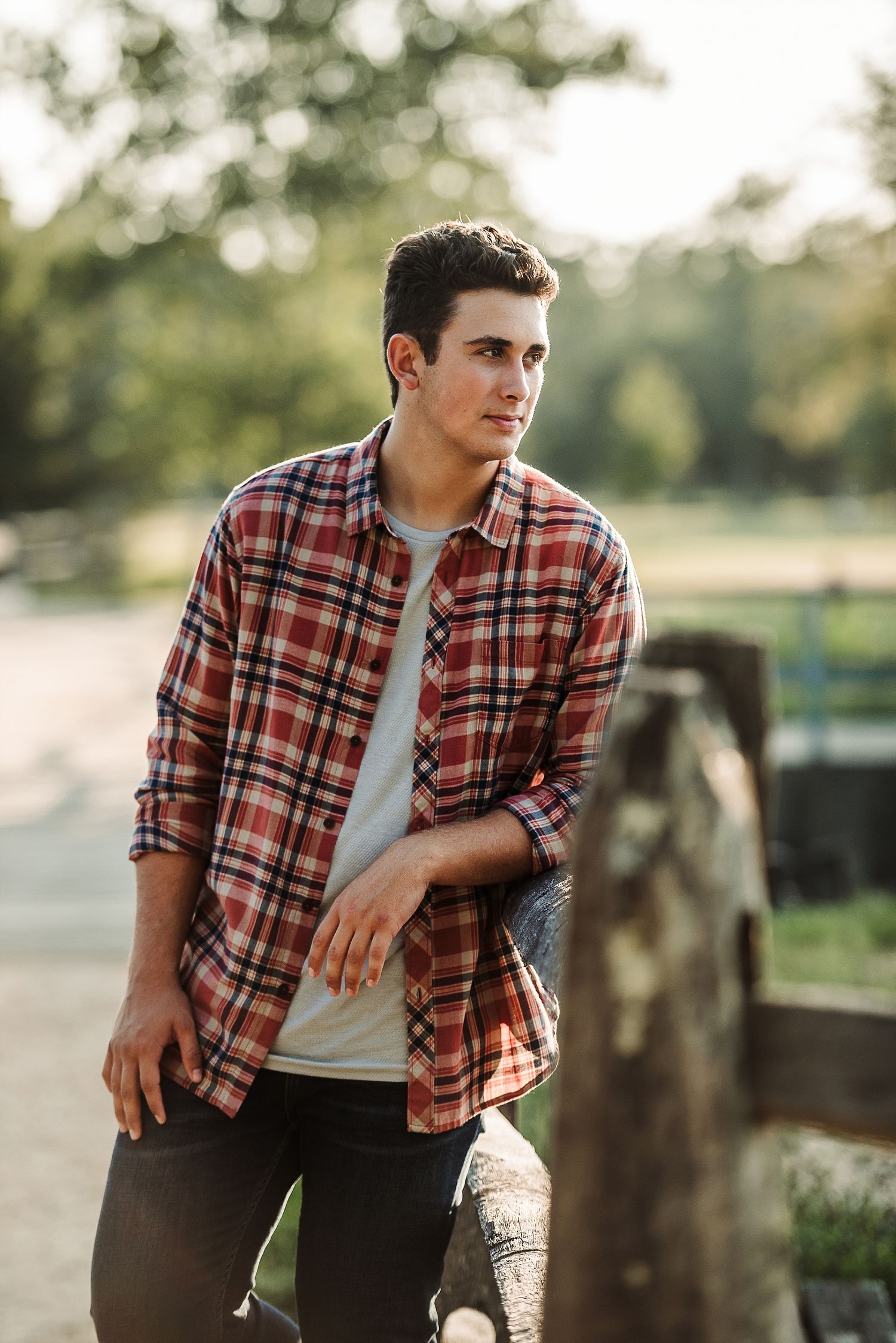 Young person in plaid flannel shirt and white t-shirt leaning against wooden fence post at sunset.