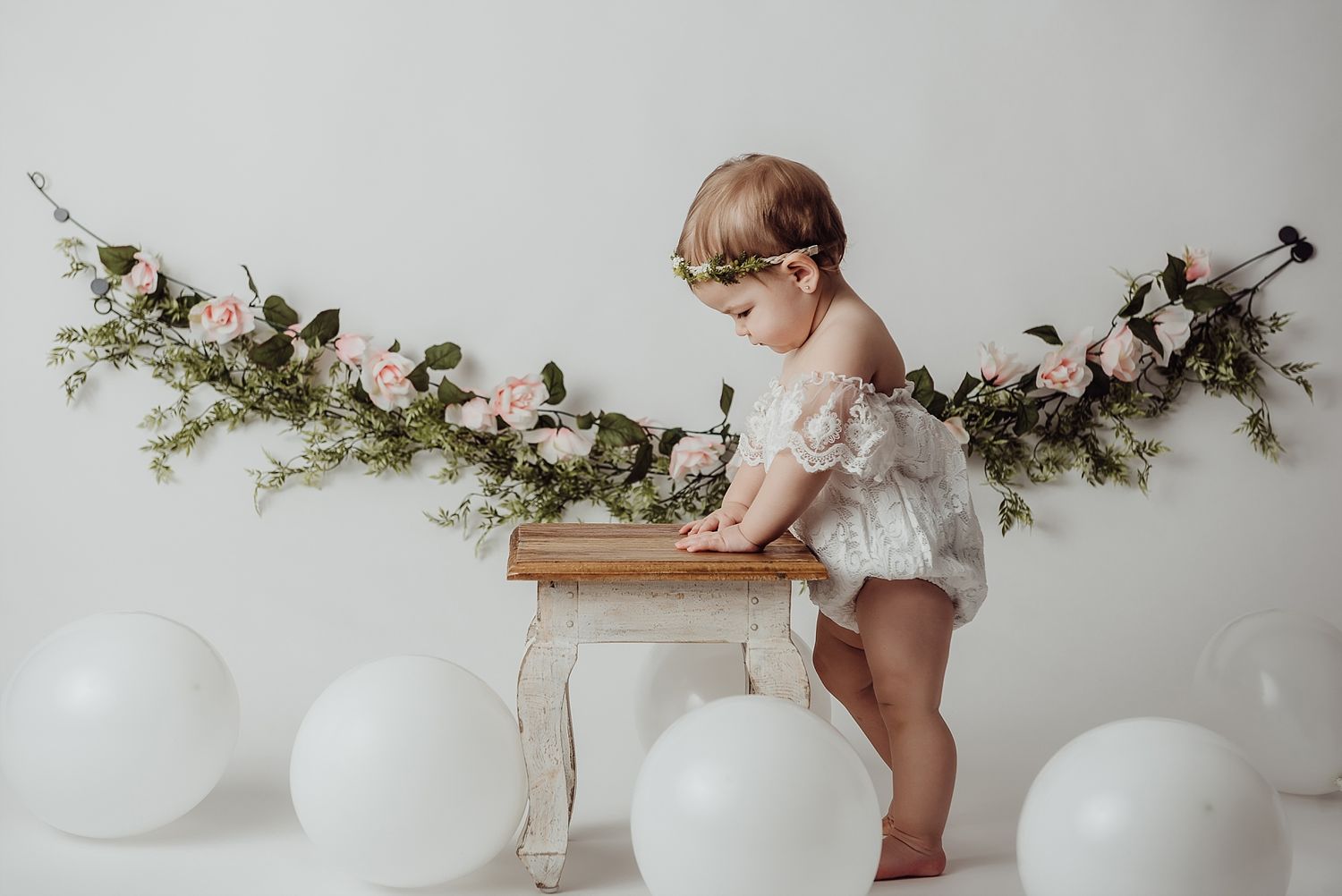 Small child in white dress stands near decorative floral garland and white balloons.