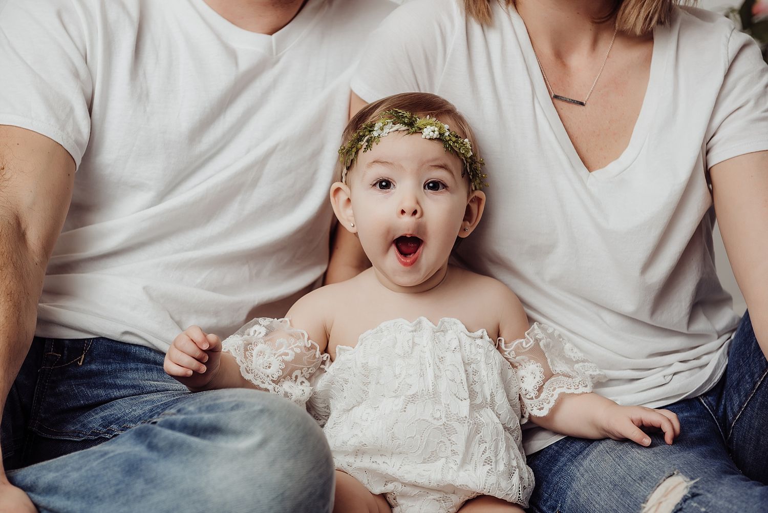 Baby in white lace dress with crown sitting between parents in matching white shirts.