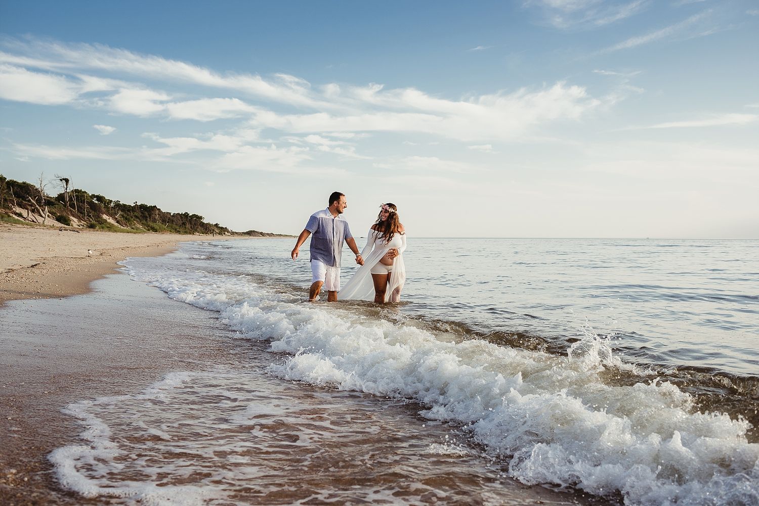 Two people walk hand in hand along a beach shoreline at sunset as waves crash onto the sand.