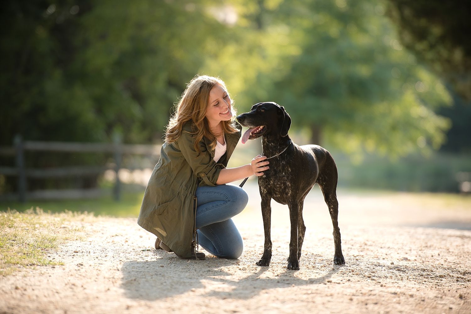 Woman in brown jacket kneels down to pet black Great Dane dog on dirt path.
