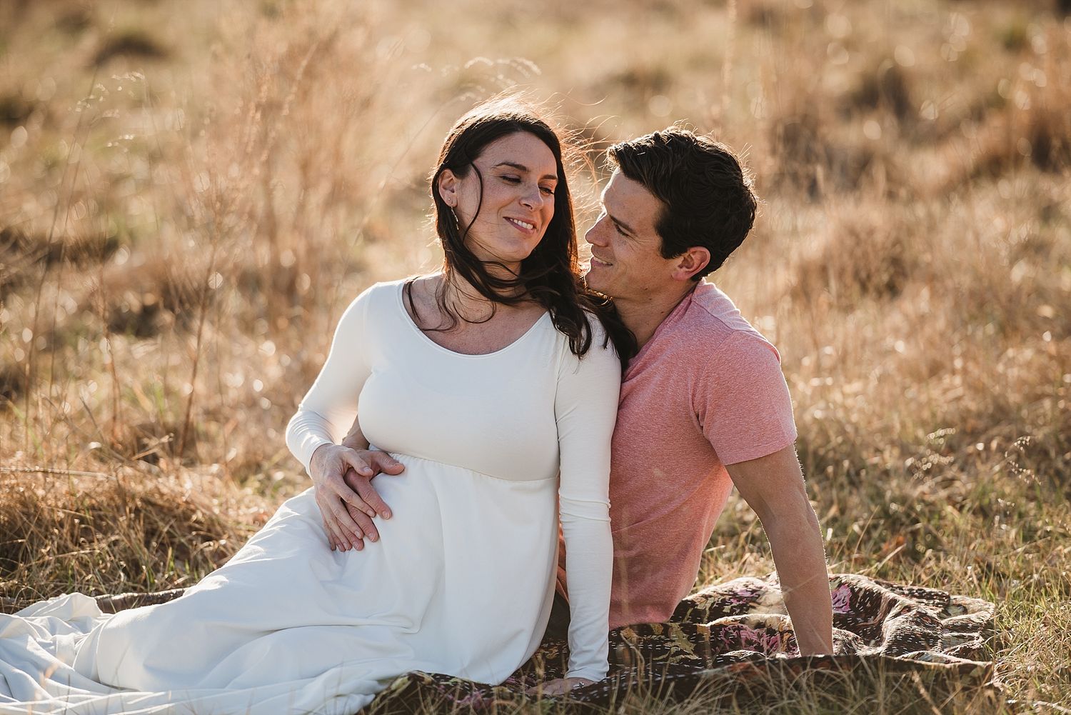 Couple sits intimately together in golden field during sunset maternity photo session.