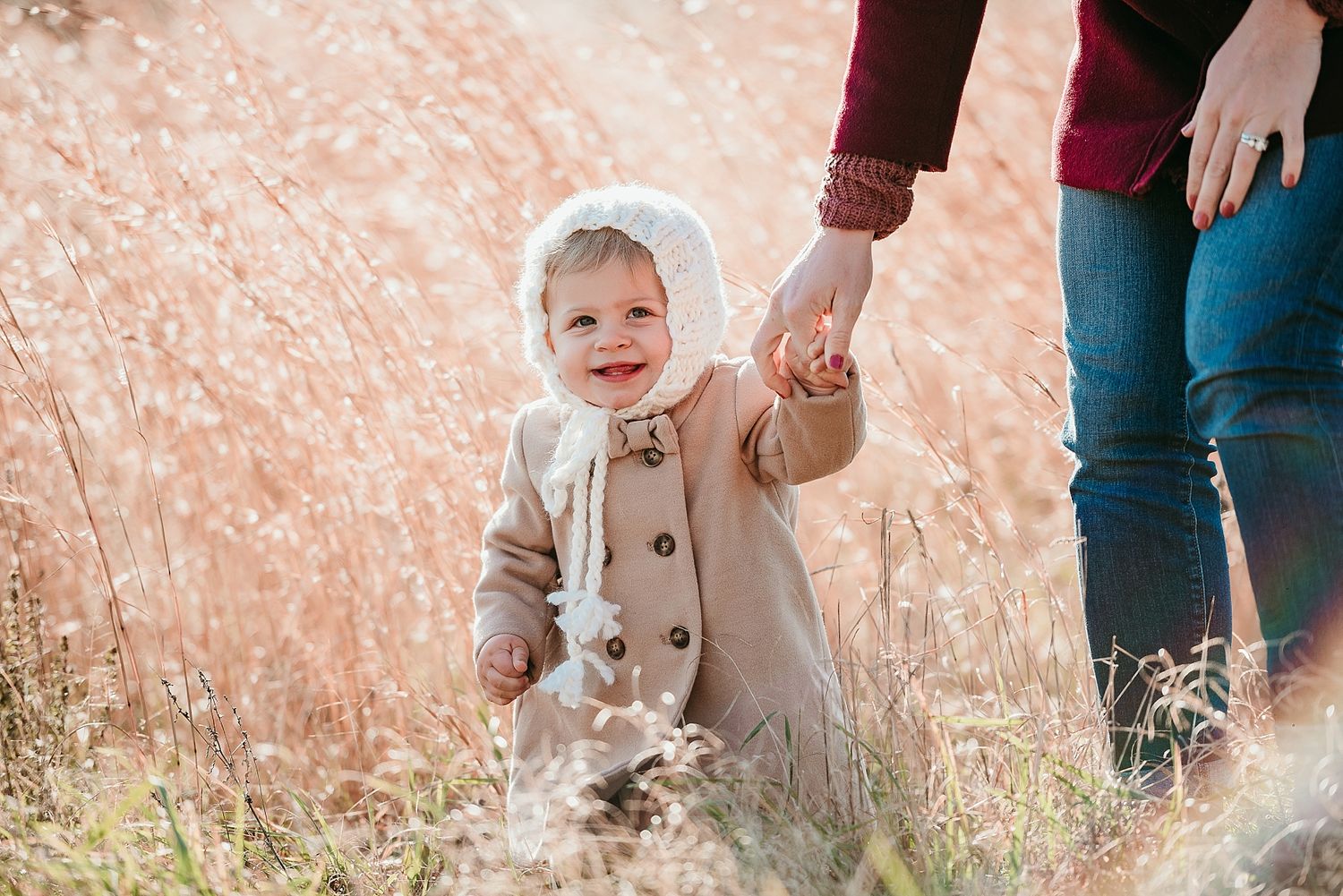 A toddler in a beige coat walks through tall grass while holding hands with someone.