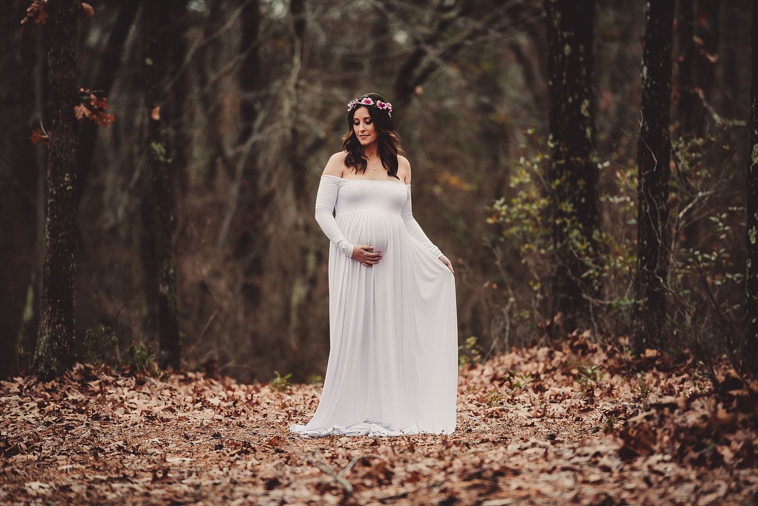 A pregnant woman in a flowing white dress stands gracefully among fallen autumn leaves in the woods.