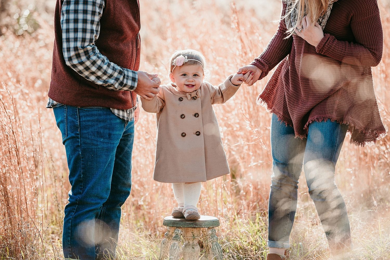A toddler in a coat takes first steps while holding hands in a field of autumn grass.
