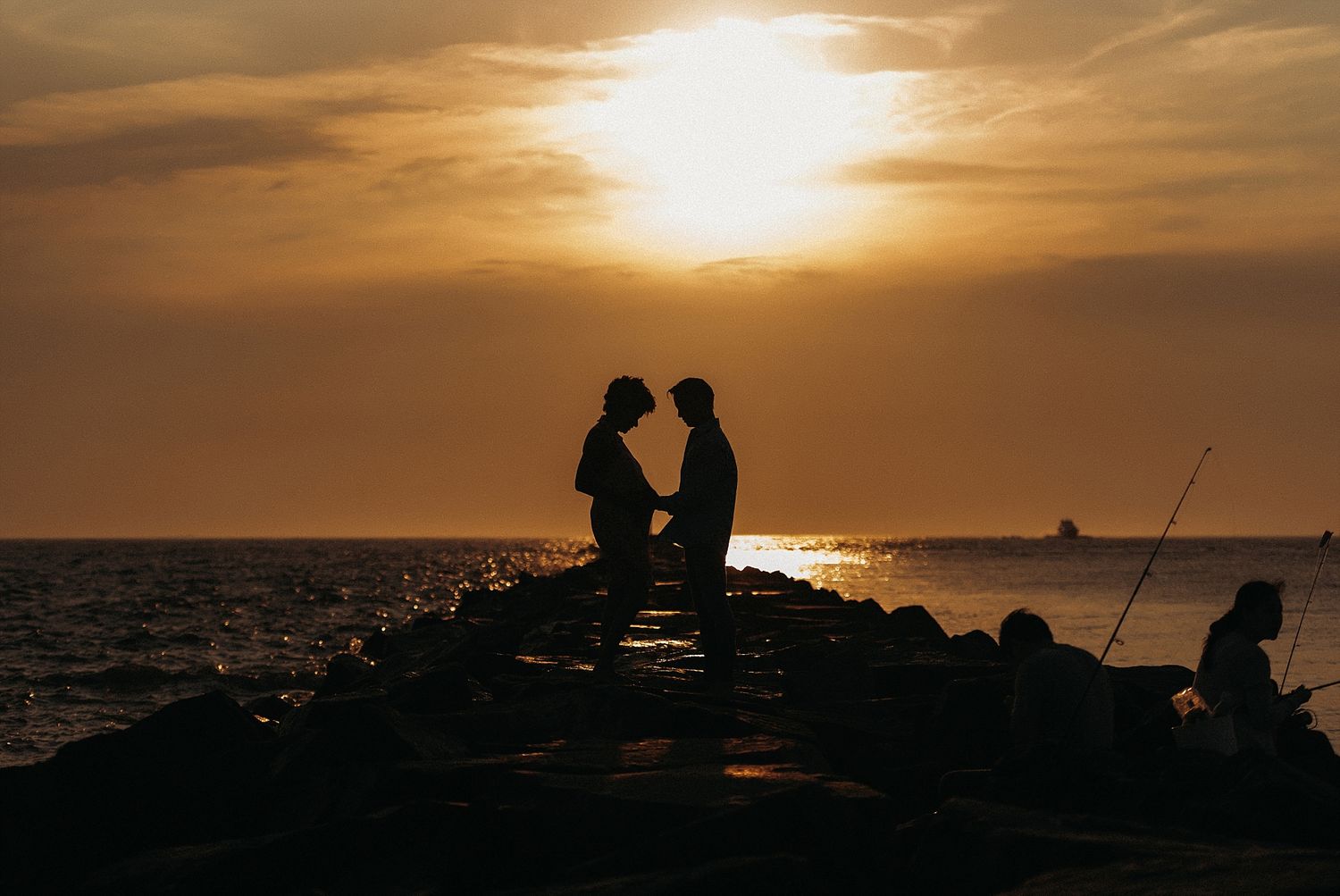 Silhouettes on rocky pier during dramatic orange sunset over water.