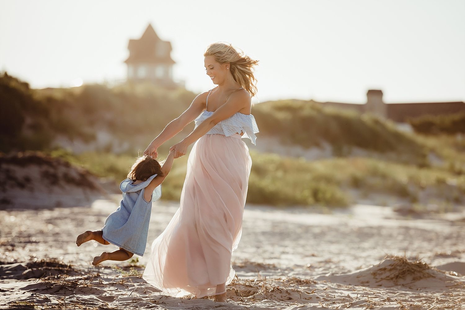 Mother and child play together on a beach at sunset with a lighthouse in the background.