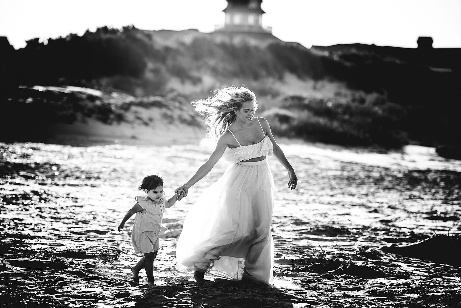 Black and white photo of two people playing on beach near lighthouse.