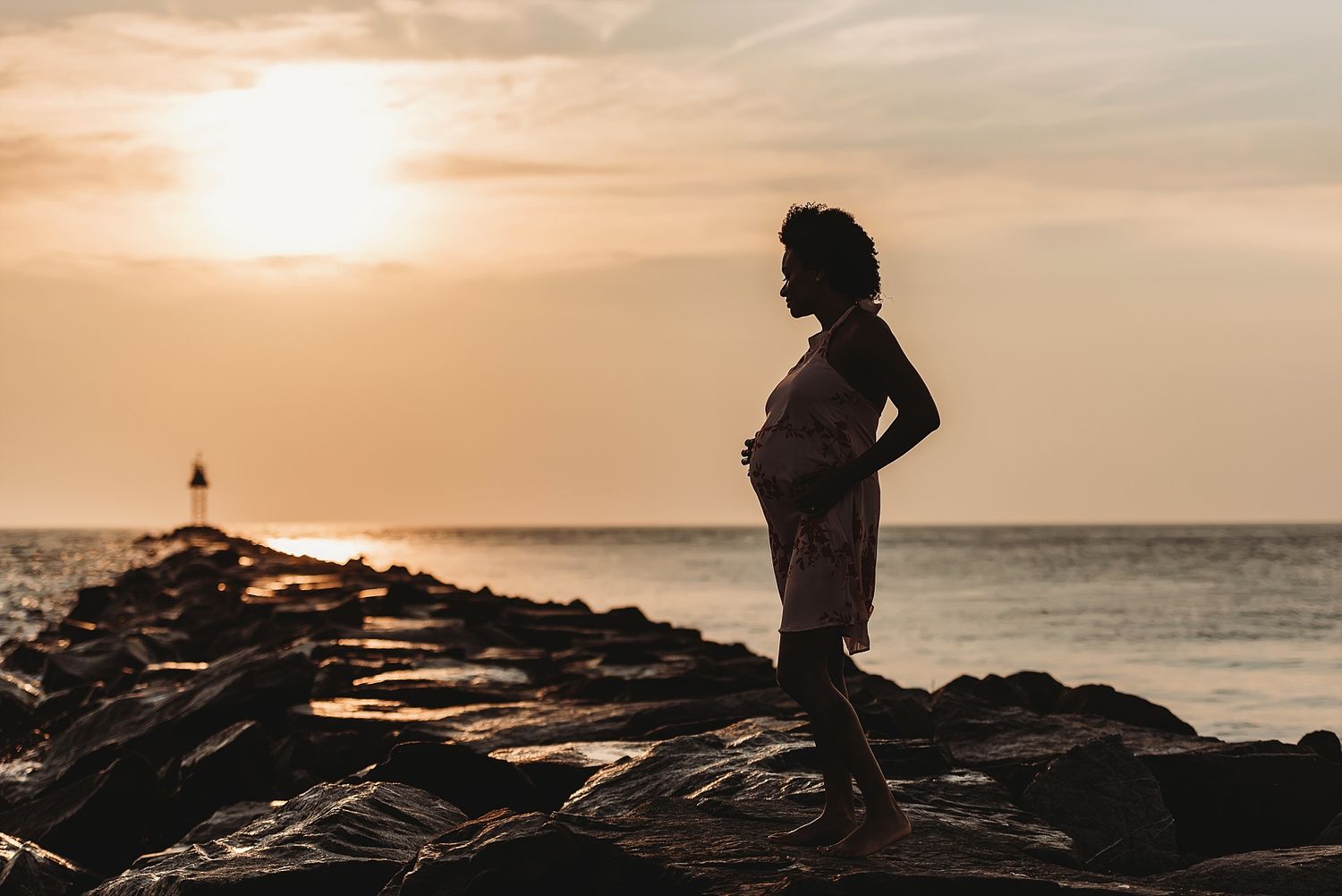 Silhouette of pregnant woman standing on rocky shore at sunset.