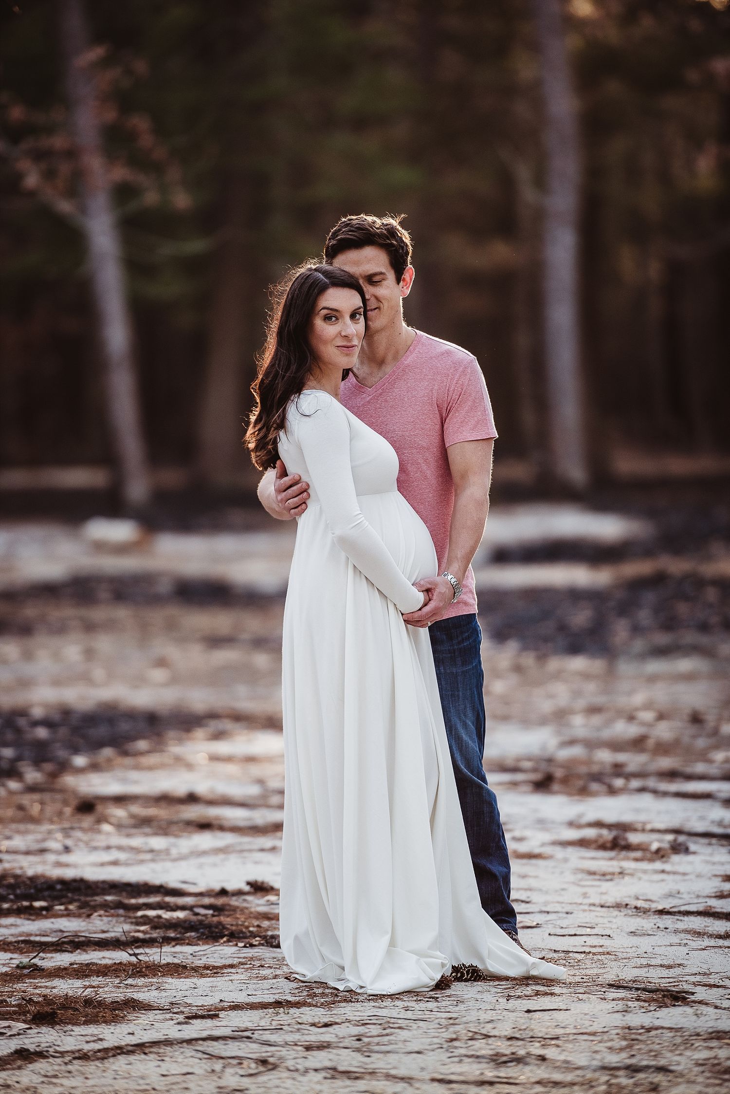 Romantic maternity photo of couple embracing in forest setting with woman in flowing white dress.