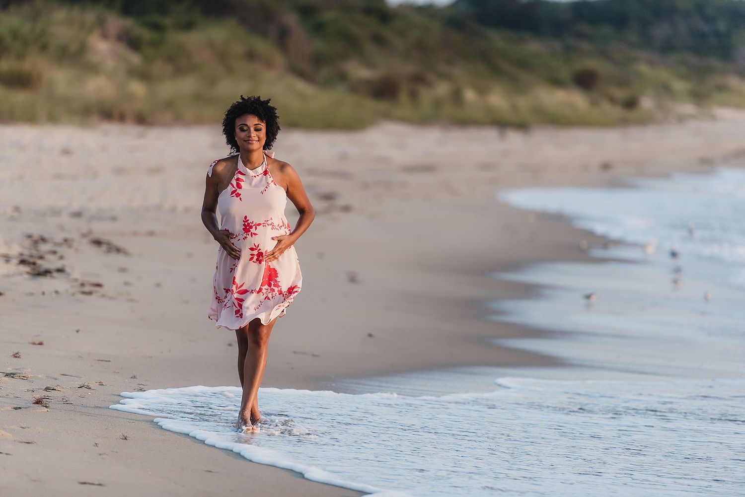 Woman in floral dress walking along beach shoreline.