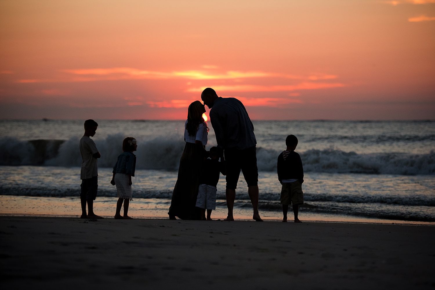 Silhouettes of family members gathered on beach at sunset with waves in background.