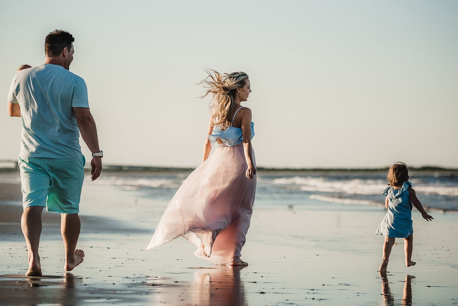 Family walking along beach at sunset with flowing dress catching ocean breeze.