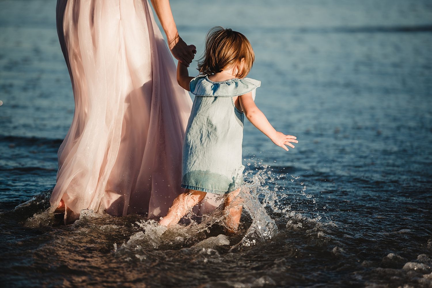 Child in blue dress splashing in ocean waves beside figure in pink dress.