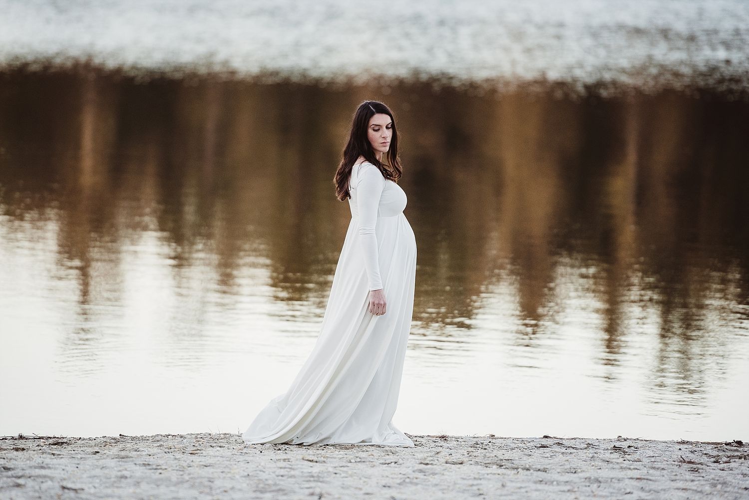 Silhouette of pregnant woman in white dress standing by still lake.