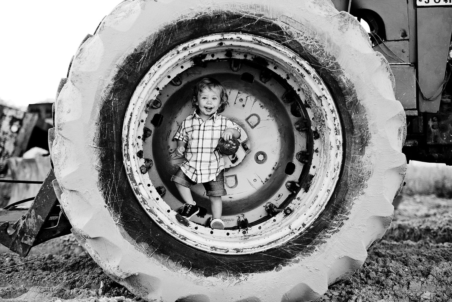 A small child sits inside a large tractor wheel in a black and white photograph.