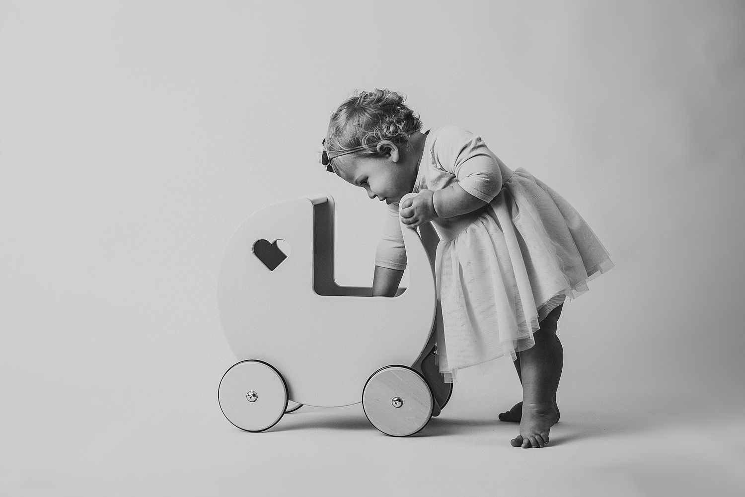 A toddler in a light dress pushes a vintage toy wagon in black and white.