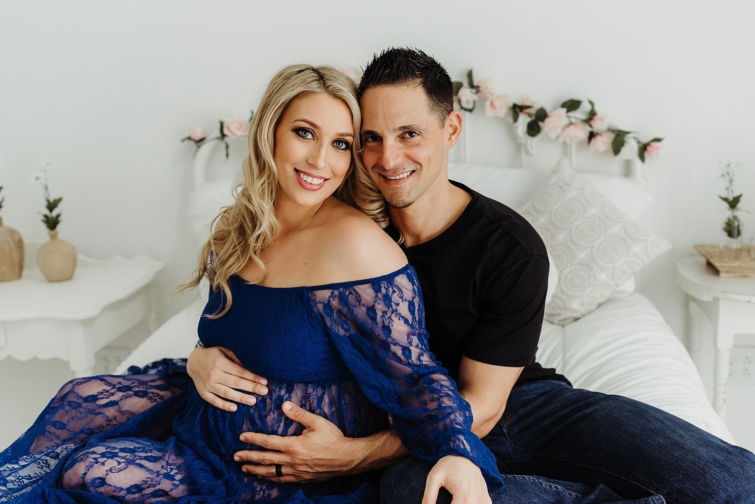 A couple poses together on a white bed with floral decorations.