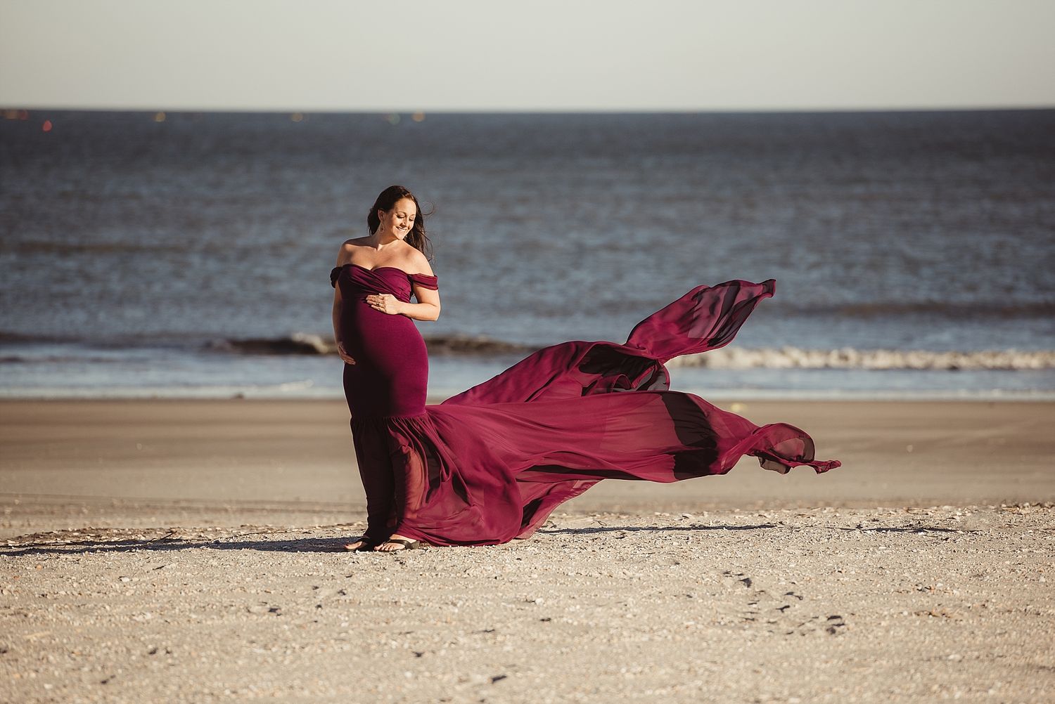 A flowing burgundy dress catches the ocean breeze as a figure stands on a sandy beach at sunset.