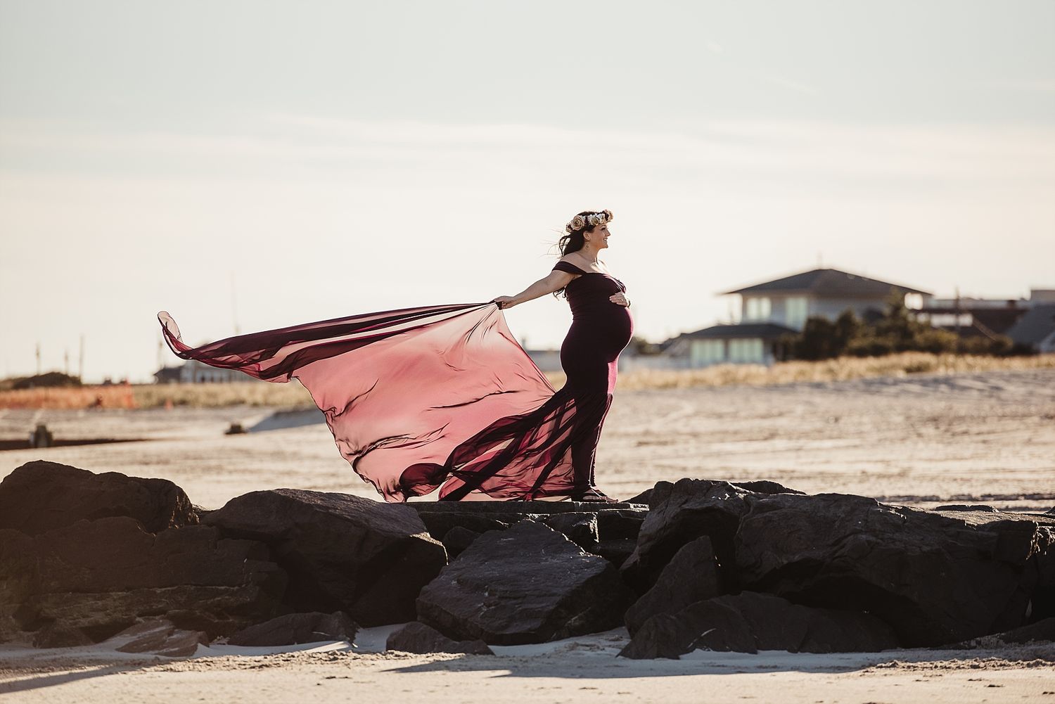A flowing pink dress billows dramatically on rocky coastal terrain at sunset.