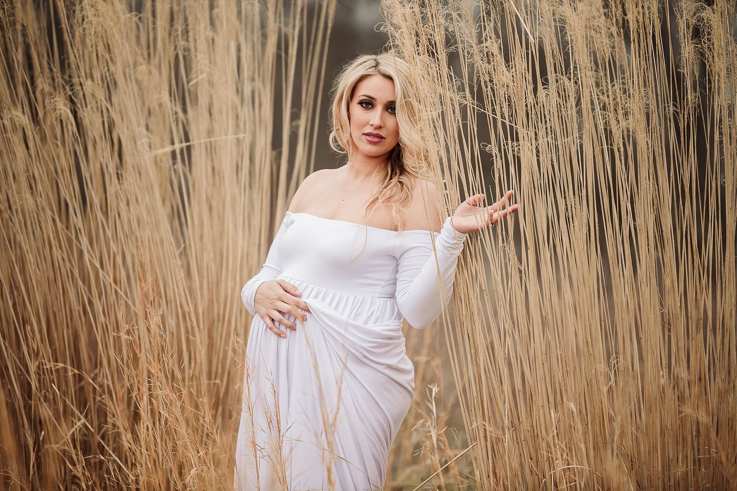 Figure in white dress poses elegantly among tall dried reeds in nature.