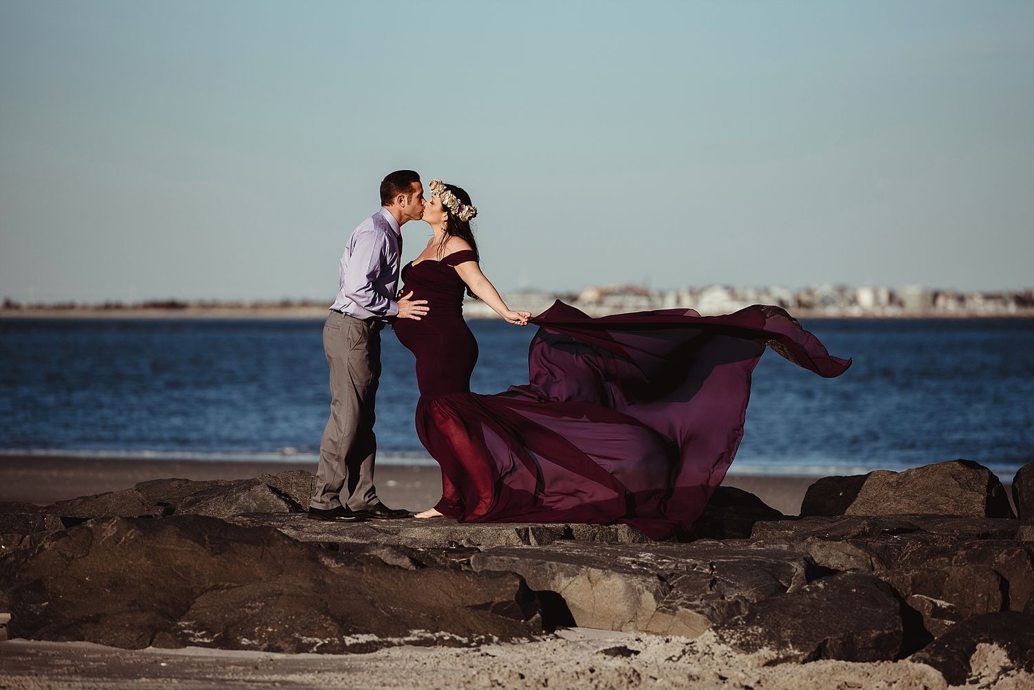 A burgundy dress flows dramatically as couple embraces on rocky coastal shore.