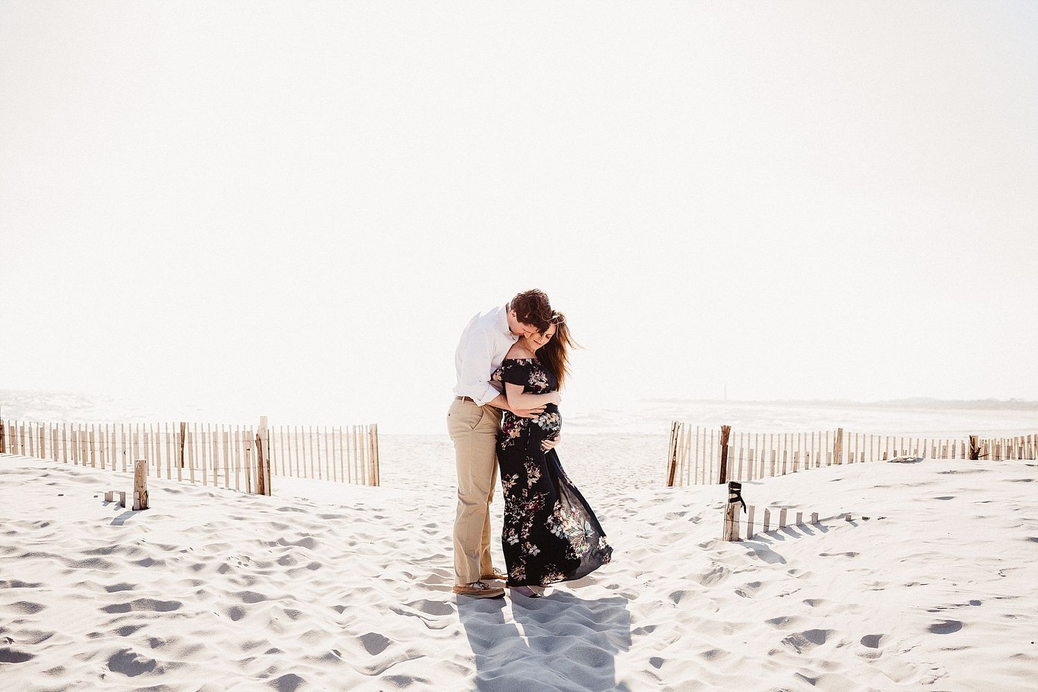 Couple embracing on sandy beach with wooden fence in background.
