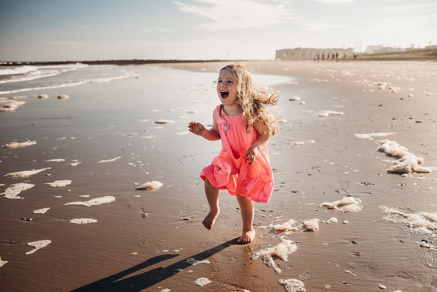A young child in a coral dress dances joyfully on a beach with seashells scattered around at sunset.