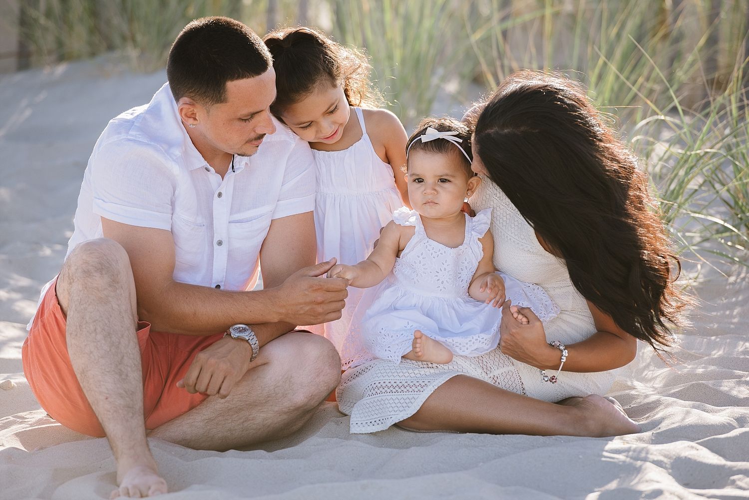 Family sits together on sandy beach dunes wearing coordinated white and coral outfits.