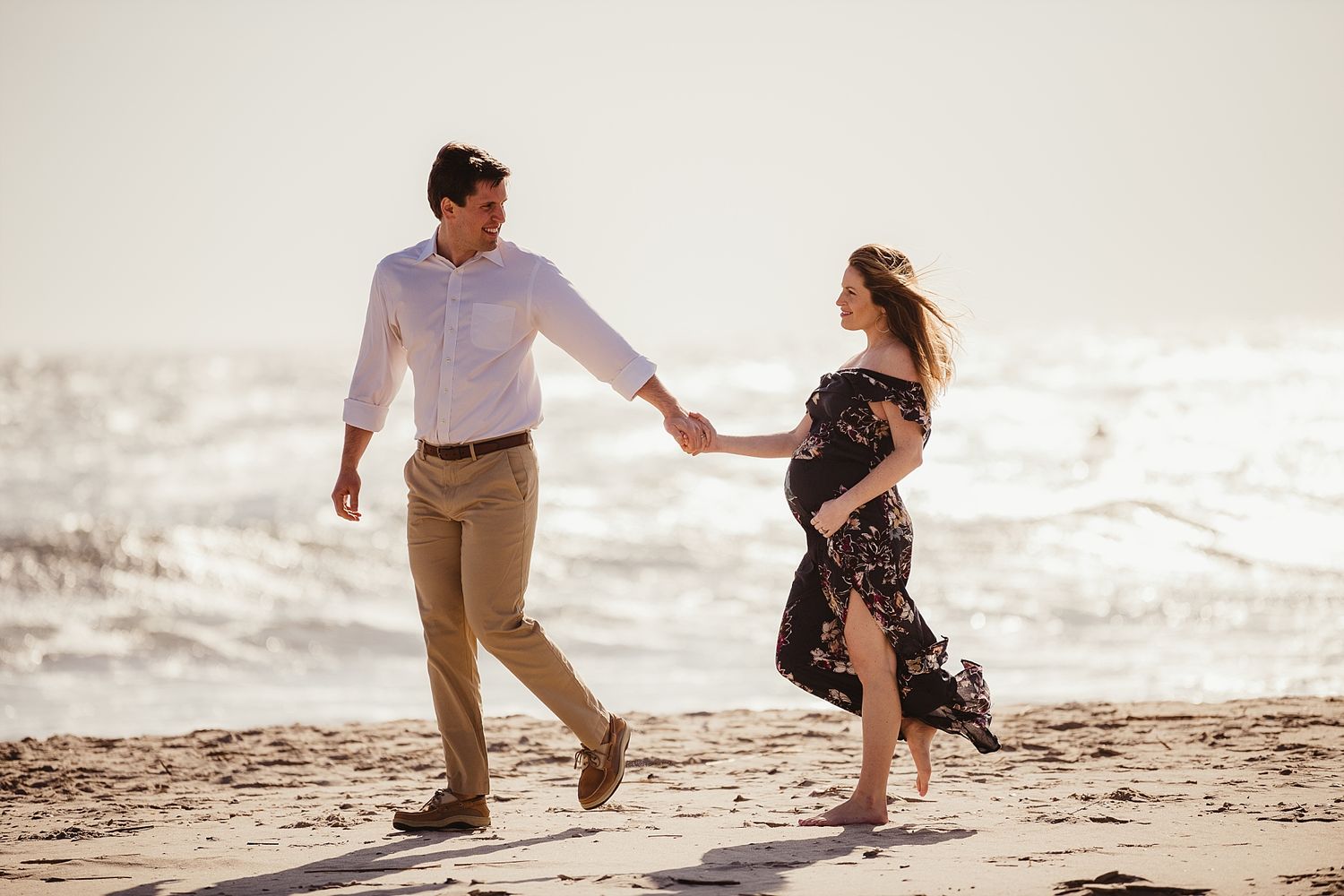 Couple holds hands while dancing together on beach at sunset.