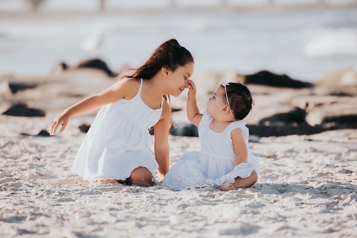 Two figures in white dresses share a sweet moment while sitting together on sandy beach.