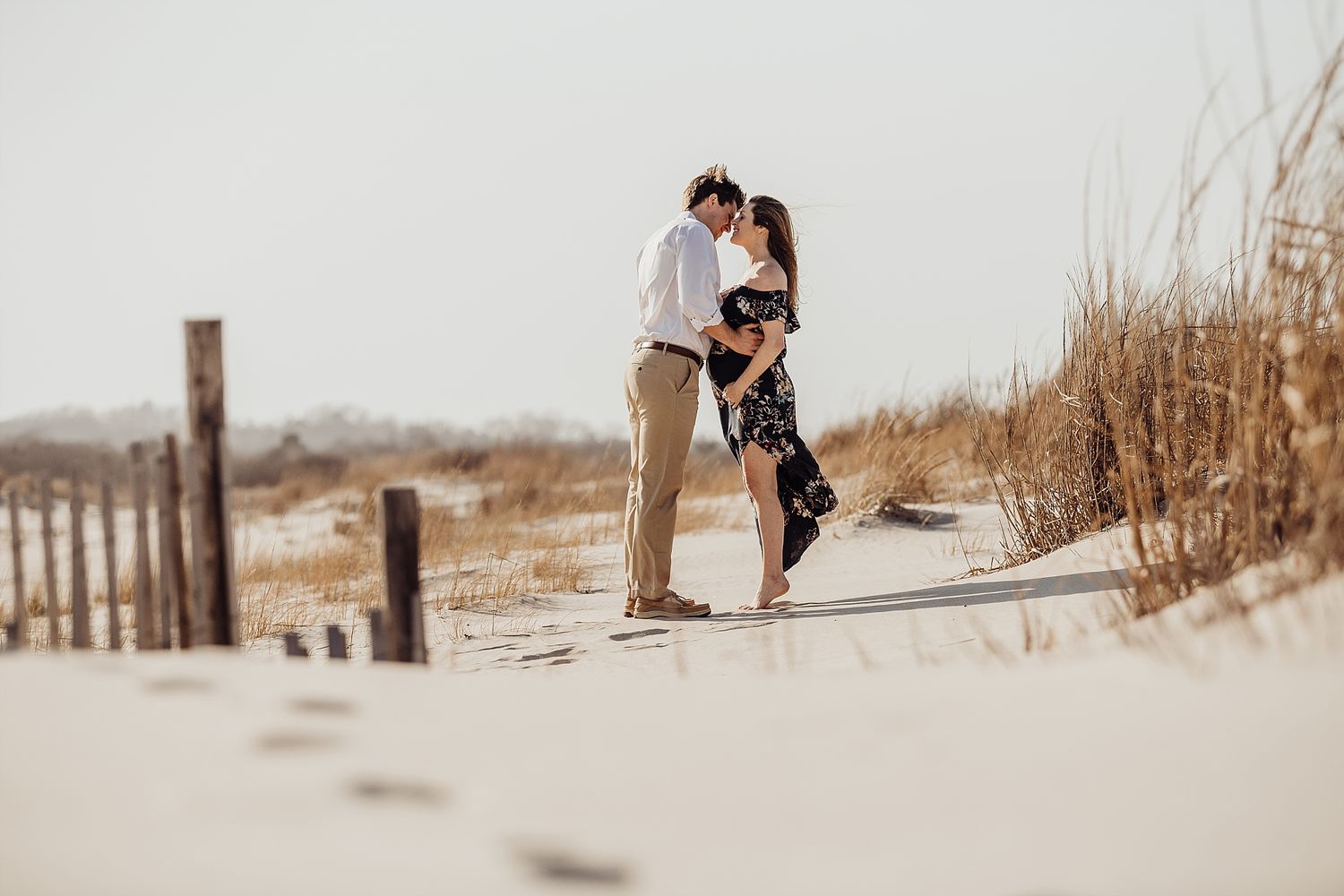 Romantic couple embraces on sandy beach path with sea grass in background.