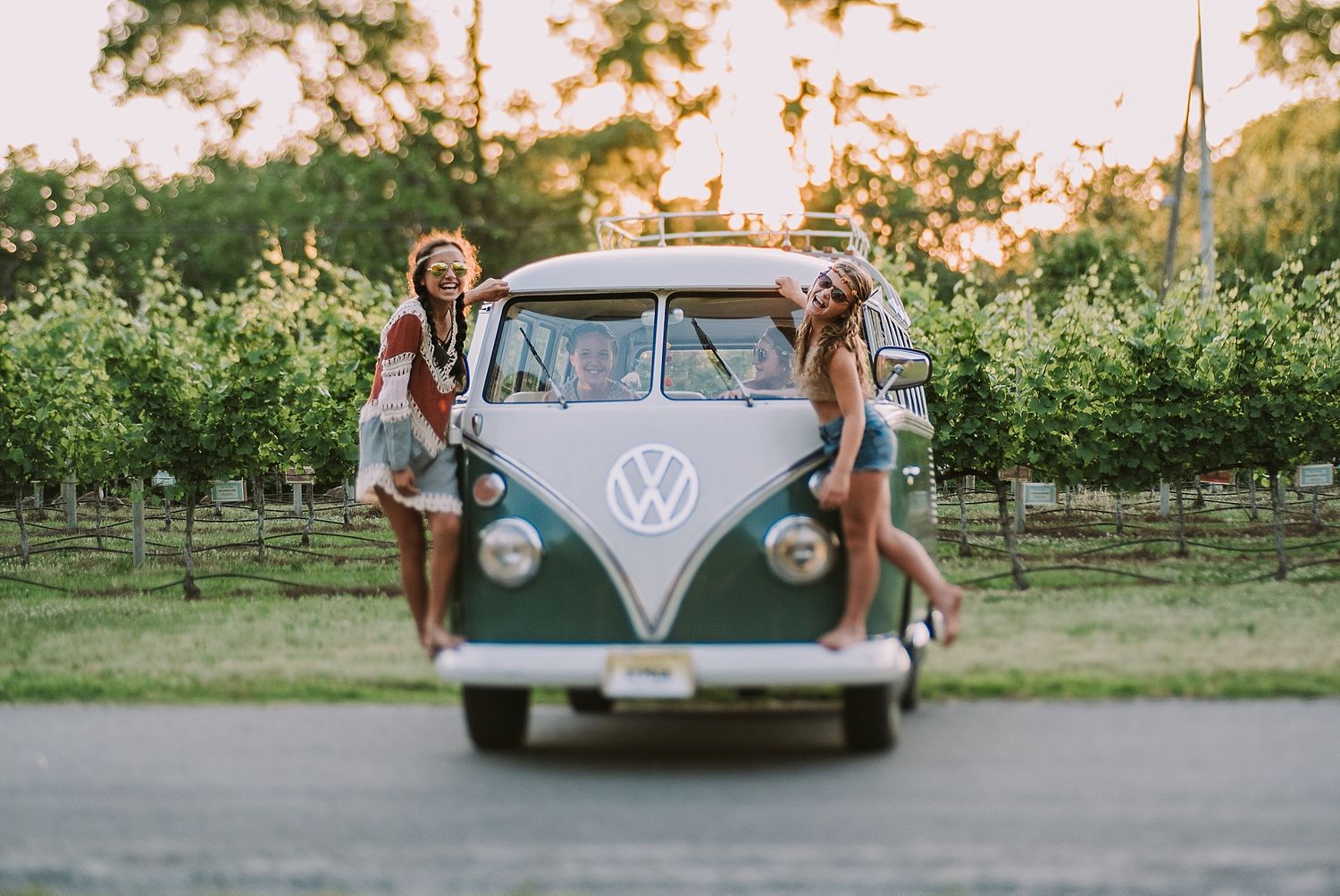 Two people pose with a classic teal Volkswagen bus in a vineyard at sunset.