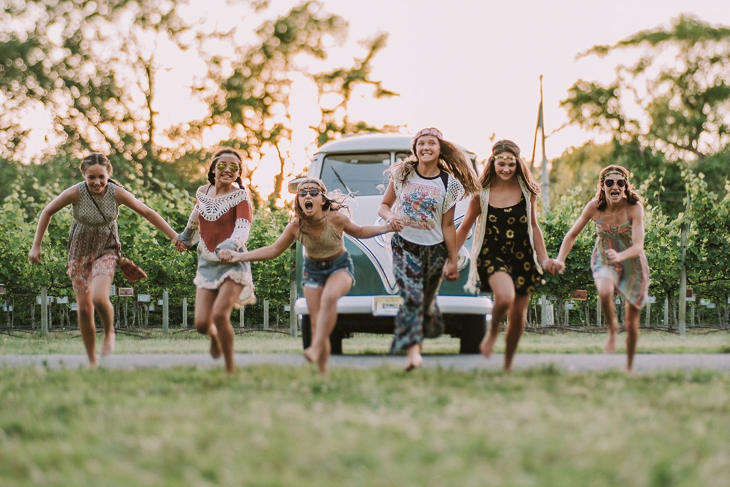 A joyful group runs and jumps through a grassy field at sunset with a vintage van in the background.