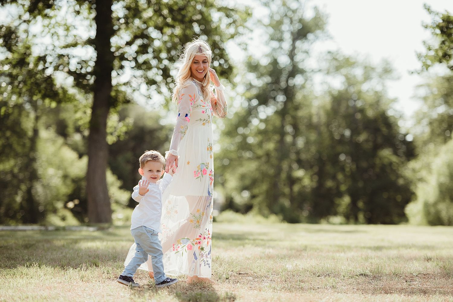 A flowing white dress moves in the breeze as a toddler walks through a sunlit park.