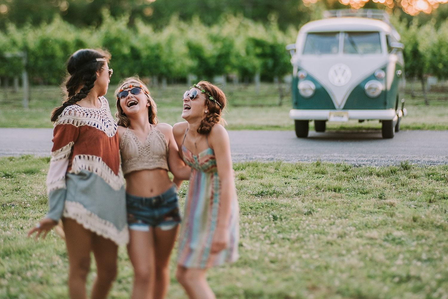 Three friends laugh together in summer outfits with a vintage VW bus visible behind them.