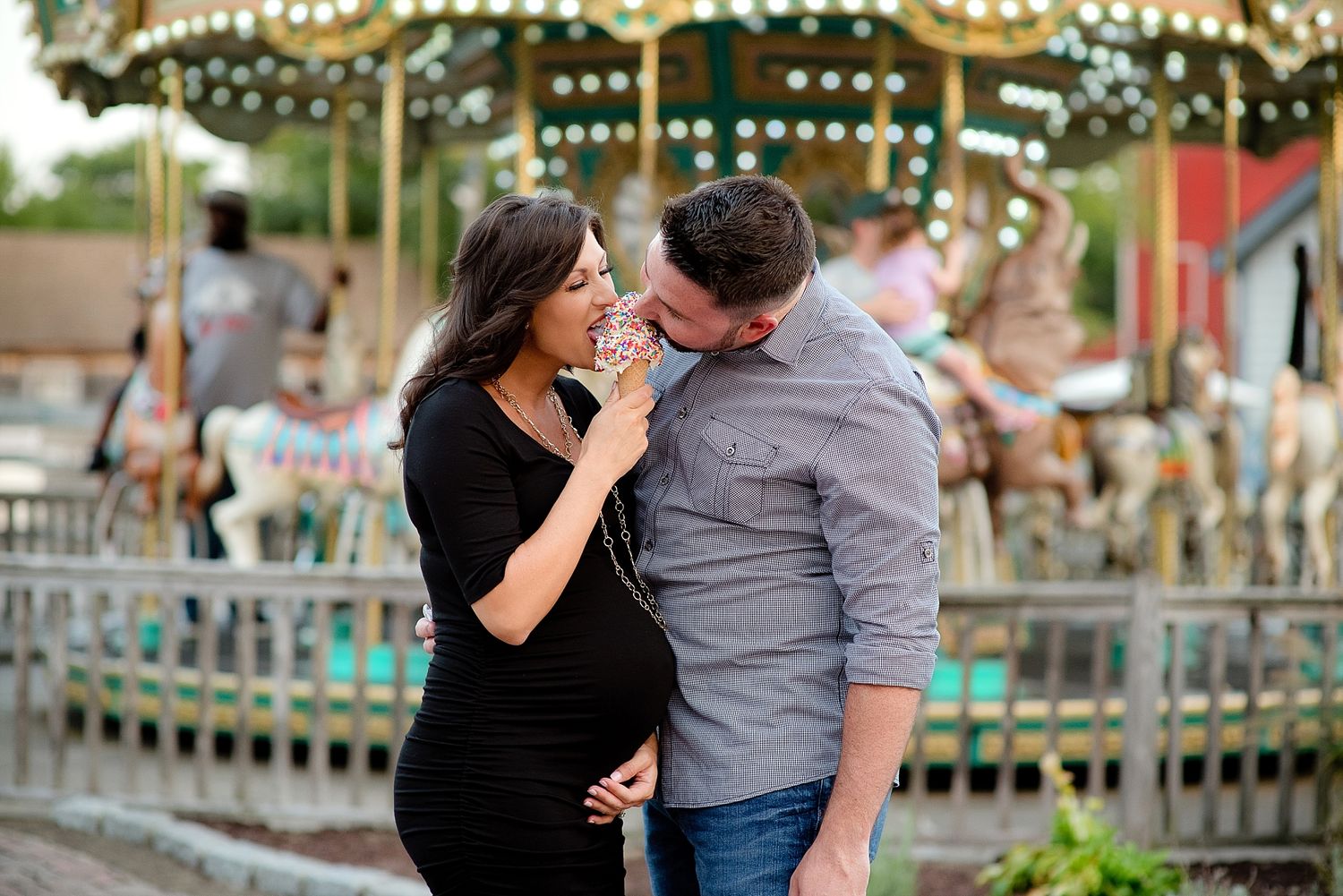 A couple shares an intimate moment in front of a carousel with twinkling lights.