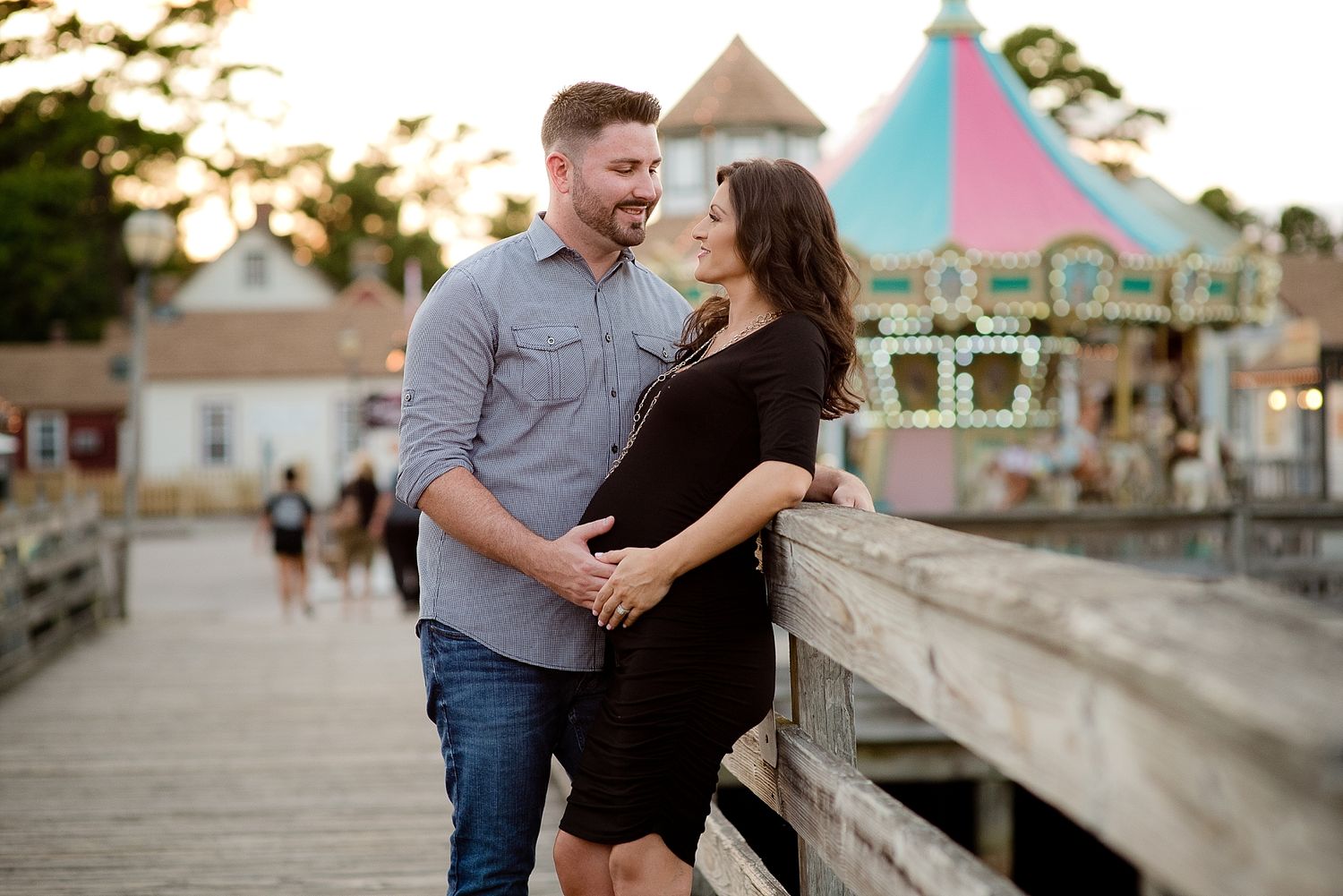 A couple shares a romantic moment on a wooden pier with a colorful carnival backdrop.