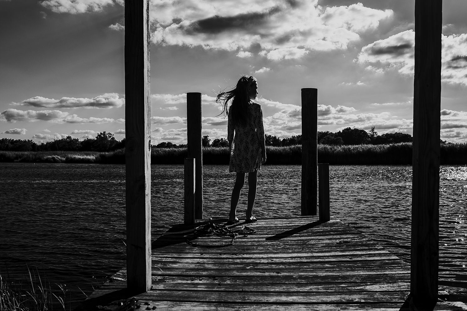 Silhouette of person standing on wooden dock at sunset over water.