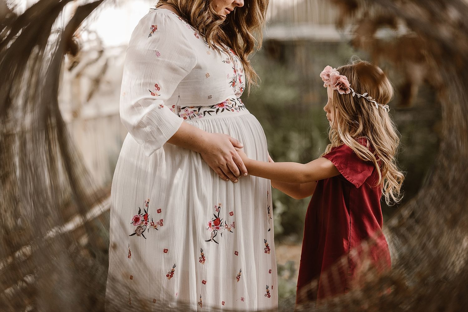 A touching maternity photo shows a floral dress-clad pregnant figure holding hands with a small person in red velvet.