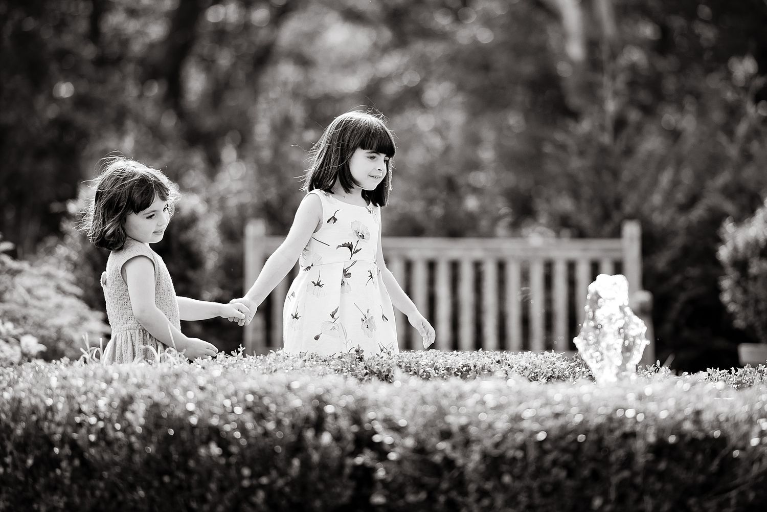 Two young girls in dresses walking together in a garden with a wooden bench in soft black and white.
