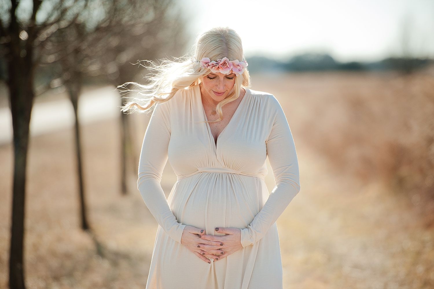 Maternity portrait in white dress with sunlight streaming through trees in winter setting.