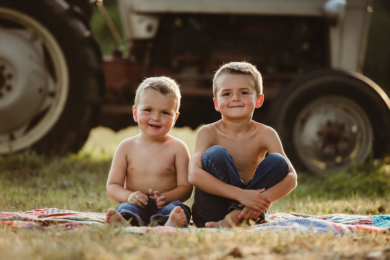Two children sit together on a blanket near a tractor in a sunny field.