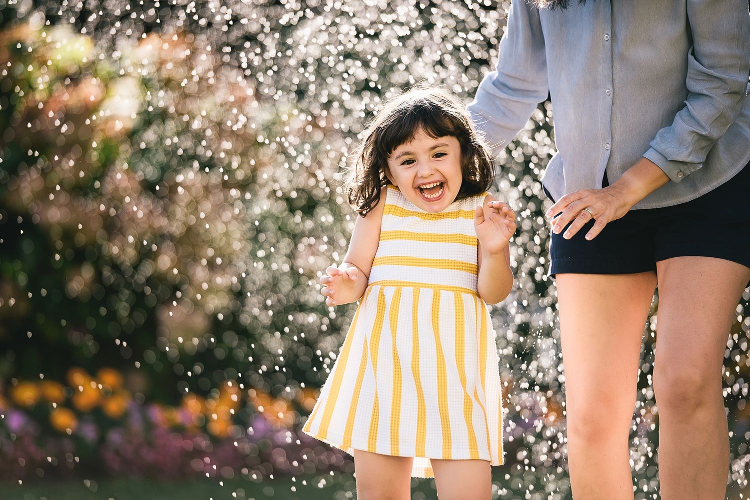 A child in a yellow striped dress enjoying water sprinklers in a garden.