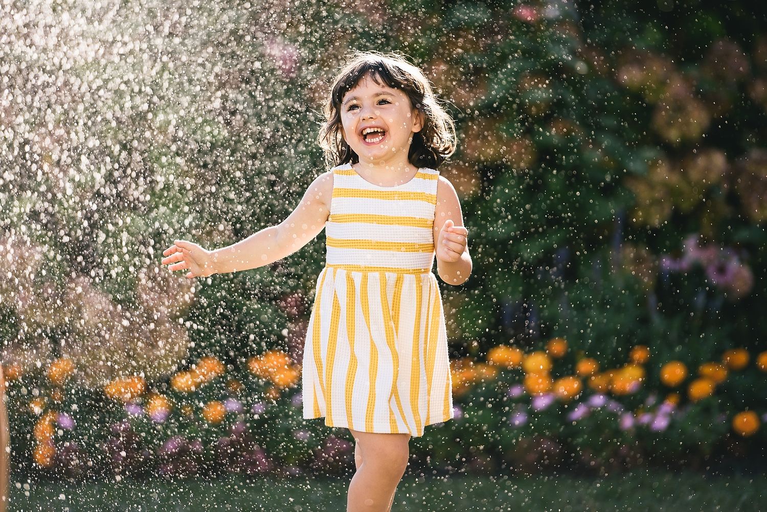 A child in a yellow dress laughing while playing in water sprinklers outdoors.