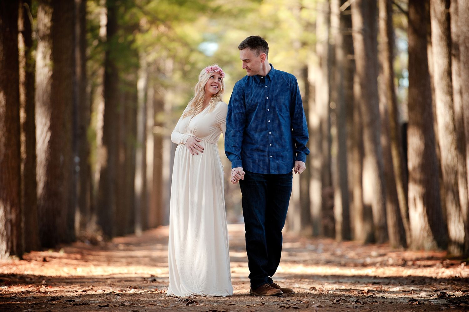 A couple in formal attire walks together through a forest pathway lined with tall trees.