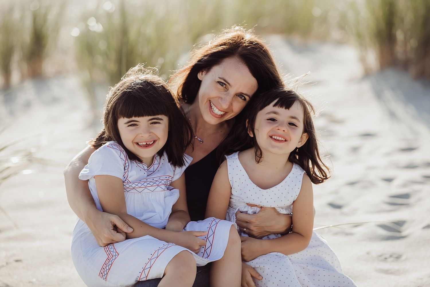 Three people wearing white dresses share joyful moments together on a sunny beach.