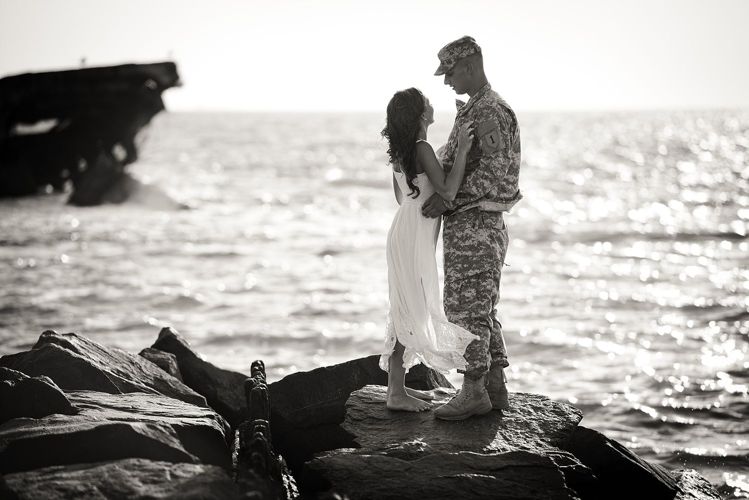 A silhouetted military service member and companion share a moment on coastal rocks at sunset.